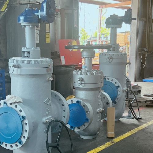 Three grey industrial gate valves with blue flange covers and blue gear operators in a workshop